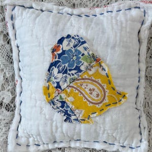 May include: A small, square white pillow with a hand-stitched bird applique. The bird is made of floral and paisley patterned fabric in blue, yellow, and orange. The pillow has blue and red stitching around the edges.
