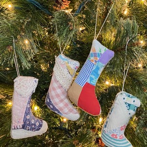 May include: Four fabric Christmas stocking ornaments hanging from strings. Each stocking features a unique patchwork design with various patterns including gingham, floral, and geometric prints in colors like red, blue, pink, and white, accented with lace.