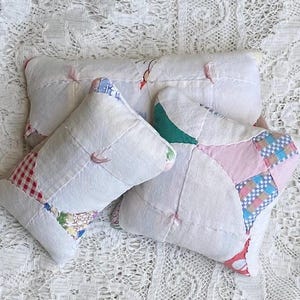 May include: Three small, handmade pillows with white fabric and patchwork accents. The patchwork includes red gingham, floral, and pastel patterns. Each pillow is stitched with pink thread and has a soft, vintage aesthetic.
