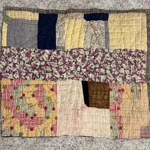 May include: A patchwork quilt featuring a variety of fabric squares in yellow, navy blue, brown, and cream, accented by floral patterns in purple and green, and striped sections with red circular motifs, all held together by visible hand-stitched quilting.