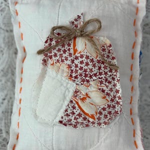 May include: A handmade, quilted pillow with a white background and an appliqued pear shape. The pear is made of fabric with a floral pattern in red and orange. The pillow is trimmed with orange stitching and tied with twine.