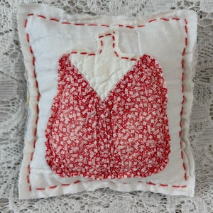 May include: A handmade, square white pillow with a red floral pattern applique. The applique is in the shape of a dress. The pillow has red stitching around the edges and the applique. The pillow is on a white lace surface.