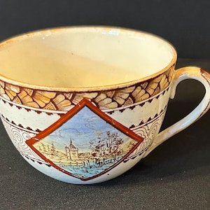May include: Cream-coloured teacup with a brown floral pattern around the rim and a diamond-shaped image of a building and landscape. The handle and rim have a brown trim.