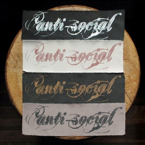 May include: Four fabric patches with the text "Anti-Social" written in a stylized font. The patches are in different colors: white, pink, gold, and black.