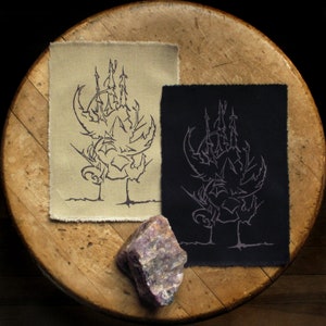 以下が含まれることがあります： ベージュと黒の布地に、スタイリッシュな城の白黒のデザインが刺繍された2つのパッチ。パッチは、中央に紫色の石が置かれた木製の表面に置かれています。