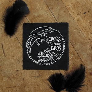 May include: Black square patch featuring a white crow graphic, the text "Crows Before Bros," and "Support Your Local Murder." Black feathers are arranged around the patch on a wooden surface.