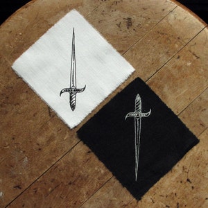Könnte beinhalten: Zwei gestickte Patches mit einem stilisierten Dolch mit floralem Design. Ein Patch ist weiß mit schwarzer Stickerei, der andere ist schwarz mit weißer Stickerei.