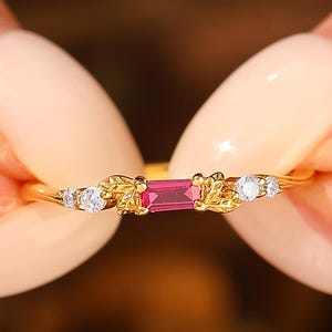 May include: A gold ring featuring a rectangular, vibrant pink gemstone at the center, flanked by small, clear gemstones and leaf-like designs. The ring is held between two hands, showcasing its delicate details.
