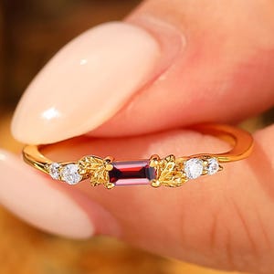 May include: A gold ring featuring a rectangular, deep red gemstone at the center, flanked by smaller white gemstones and leaf-like designs. The ring is held up by a person's fingers.