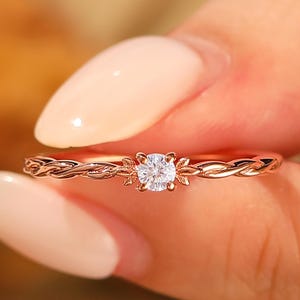 May include: A delicate rose gold ring with a small, sparkling diamond centre stone. The band has a braided design with floral accents around the diamond. The ring is held up against a blurred background.