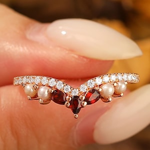 May include: A rose gold ring with a curved design. The ring features small pearls, red gemstones, and a row of small clear stones. The ring is held by a person.