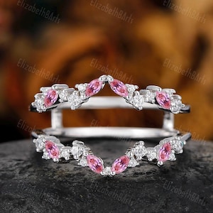 May include: A delicate silver ring enhancer with a V-shaped design. The enhancer is adorned with small, round clear diamonds and pink marquise-shaped gemstones.