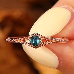 May include: A rose gold ring featuring a round, deep blue gemstone set in a hexagonal frame. The band has a delicate, beaded texture along the sides, leading to a split shank design that meets at the central stone setting.