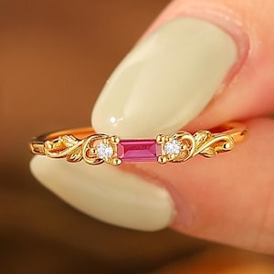 Peut inclure: Une bague en or ornée d'une pierre rose rectangulaire et de deux petites pierres rondes et transparentes. La bague présente un motif floral orné de chaque côté des pierres précieuses. La bague est tenue par une main avec du vernis à ongles vert clair.