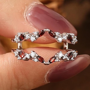May include: A silver ring guard with a floral design. The ring guard features small, round, and marquise-cut red gemstones and clear, round gemstones. The ring guard is held between two fingers.