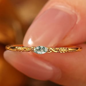 Può includere: Un delicato anello d'oro con una gemma ovale verde chiaro. L'anello presenta un intricato design floreale sulla fascia. L'anello è tenuto tra le dita di una persona.