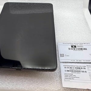 Peut inclure: Une tablette Samsung Galaxy Tab A 8.4 noire avec un écran sombre, posée sur une surface blanche. Deux étiquettes blanches avec du texte et des codes-barres sont à côté de la tablette. La tablette a un design élégant et moderne.