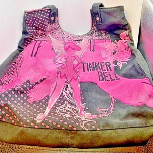 May include: A gray canvas tote bag with a pink and black design featuring Tinker Bell. The bag has a pink polka dot pattern and a pink floral design. The text "Tinker Bell" is printed in white on the bag.