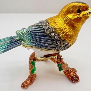 Pode incluir: Estatueta decorativa de pássaro com cabeça e corpo dourados, asas azuis e turquesas e base de ramo castanho. O pássaro é adornado com cristais transparentes. É uma peça ornamental detalhada.