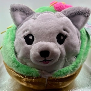 Puede incluir: Un peluche con forma de lobo gris dentro de una concha de taco. El lobo tiene ojos negros y una lengua rosa. La concha del taco es dorada y el relleno es verde. Una flor rosa está encima del taco.