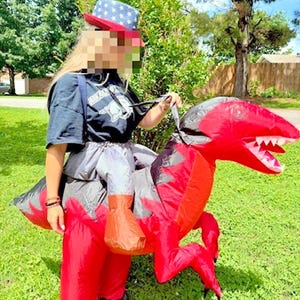 May include: An inflatable red and black dinosaur costume featuring a long tail, sharp teeth, and clawed feet. The costume is worn around the waist, creating the appearance of riding the creature. It is paired with a blue hat with white stars.