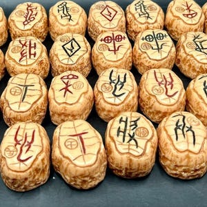 Może przedstawiać: Kolekcja trzydziestu dwóch beżowych, owalnych kamieni runicznych. Każdy kamień ma unikalny symbol w czarnym lub czerwonym tuszu. Kamienie są ułożone w równych rzędach na ciemnej powierzchni, ukazując ich kunszt.