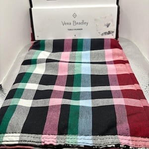 May include: A Vera Bradley table runner with a plaid design in black, white, green, pink, blue, and red. The table runner is folded and shown with its original packaging.