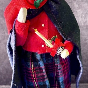 May include: A vintage Christmas carol singer figurine. The doll wears a red coat, hood, and plaid skirt. It holds a candle and a sprig of holly. The doll has a painted face and a black cape.