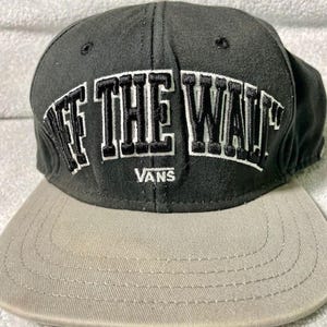 以下が含まれることがあります： 白と黒の文字で「OFF THE WALL」と書かれた黒とグレーの野球帽。ブランド名「VANS」が下に印刷されています。キャップにはフラットブリムが付いています。
