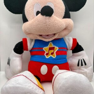 Könnte beinhalten: Ein Mickey Mouse Plüschtier mit großen schwarzen Ohren, einem beigen Gesicht und weißen Handschuhen. Die Figur trägt ein blaues Oberteil mit roten Streifen und einem gelben Stern-Emblem mit roter Musiknote sowie eine rote Hose mit zwei weißen Ovalen.