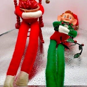 May include: Two vintage Christmas elf dolls. One elf is seated in a red chair, wearing a red outfit with white trim, a green hat, and holding a red object. The other elf is lying down, wearing a green outfit with red trim.