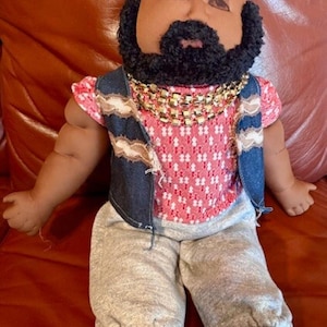 May include: A handmade doll with a brown complexion, black beard, and a mohawk-style hairstyle. The doll wears a red and white patterned shirt, a denim vest, grey sweatpants, and a gold chain necklace. The doll's feet are brown.