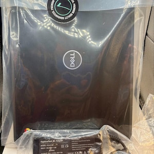 Puede incluir: Un portátil Dell negro en una funda de plástico transparente con una pegatina circular Premium Quality Guaranteed, acompañado de un adaptador de CA negro de repuesto con las especificaciones técnicas impresas en la etiqueta.