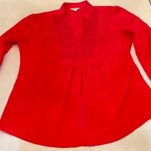 Może przedstawiać: Jasnoczerwona bluzka z długimi rękawami i dekoltem w serek. Bluzka posiada mały, haftowany detal przy dekolcie i lekko rozszerzany dół. Tkanina wydaje się lekka i odpowiednia na cieplejszą pogodę.