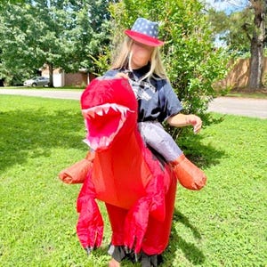 May include: An inflatable red Tyrannosaurus Rex costume with a wide open mouth and sharp white teeth. The costume has large arms and legs. A person is wearing the costume and a hat with a red brim and blue stars.