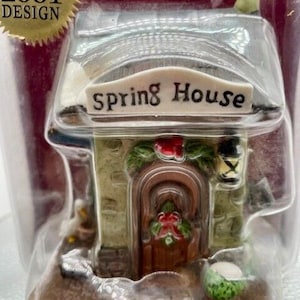 May include: A small ceramic house with a green roof and a sign that reads "Spring House". The house has a brown door with a red wreath and a small chimney. The house is sitting on a wooden base.