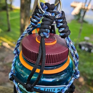 May include: A blue and black paracord bottle holder with a metal ring at the top. The bottle holder is wrapped around a clear water bottle with a maroon lid and orange accents. The bottle has several stickers on it, including one that says "This Way".