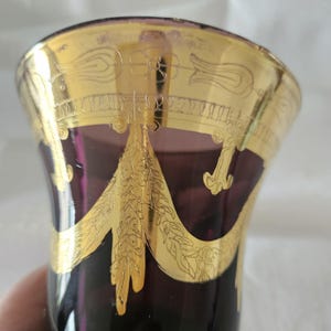 May include: A deep purple glass vessel featuring a wide, flared rim adorned with an intricate gold band. The gold overlay displays etched floral and scrollwork patterns, including stylized leaves and hanging garlands that drape down the glass.