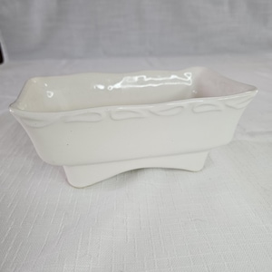 May include: A rectangular white ceramic planter with a smooth, glossy finish. The vessel features a decorative embossed pattern along the upper rim and is supported by four short, integrated feet that create a slight arch along the base.