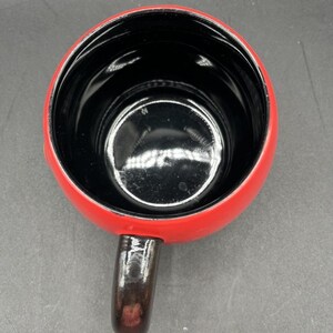 Pu&ograve; includere: Una tazza in ceramica rossa con interno nero e manico marrone scuro. La tazza &egrave; vista dall'alto, mostrando la sua forma rotonda e la finitura lucida. Il manico &egrave; attaccato al lato della tazza.