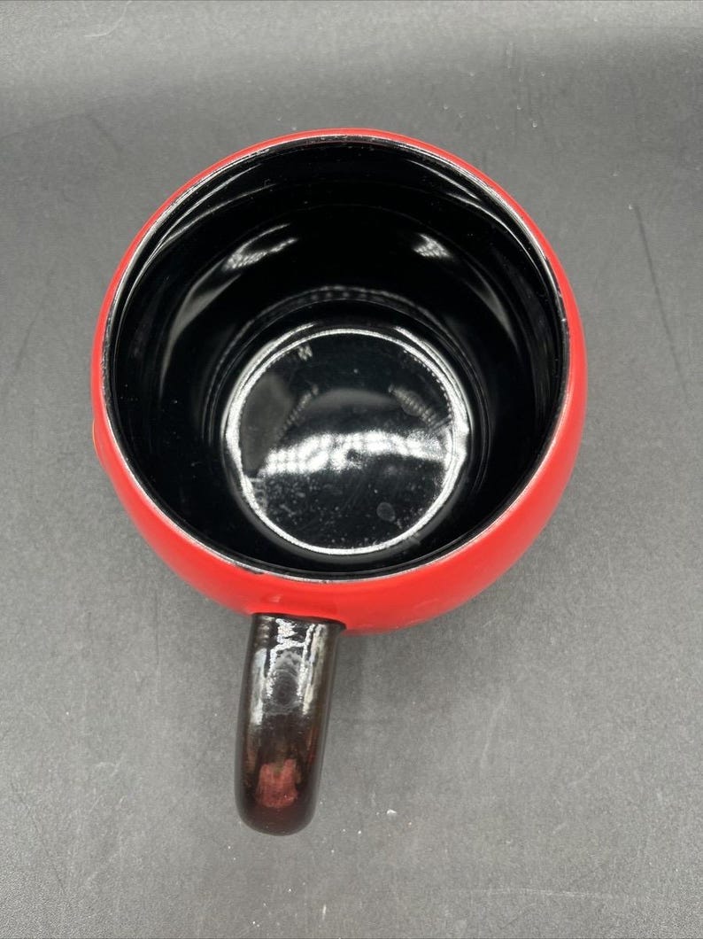 Pu&ograve; includere: Una tazza in ceramica rossa con interno nero e manico marrone scuro. La tazza &egrave; vista dall'alto, mostrando la sua forma rotonda e la finitura lucida. Il manico &egrave; attaccato al lato della tazza.