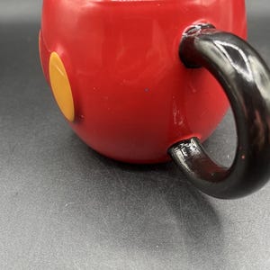 Pu&ograve; includere: Una tazza in ceramica rossa con manico e interno neri. La tazza presenta due cerchi gialli sul lato, che ricordano un personaggio dei cartoni animati. La tazza &egrave; su una superficie grigia.