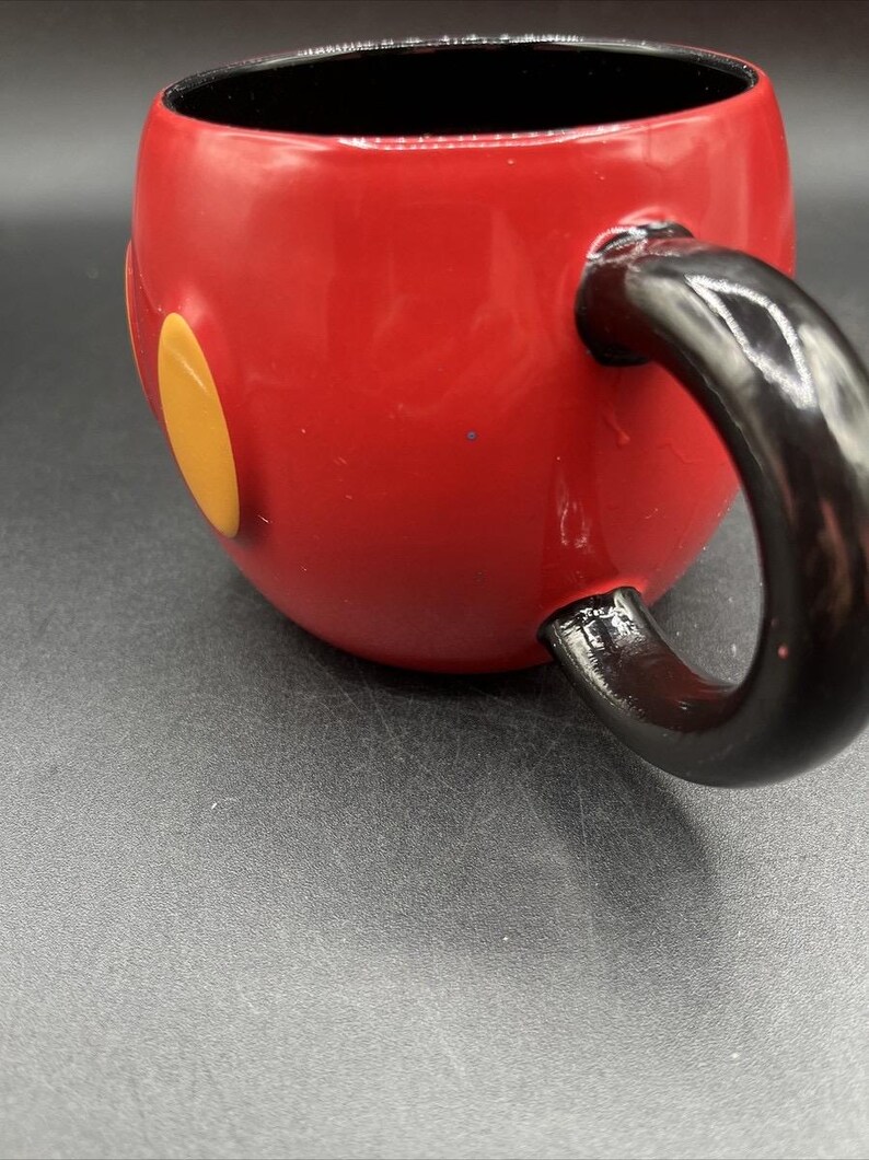 Pu&ograve; includere: Una tazza in ceramica rossa con manico e interno neri. La tazza presenta due cerchi gialli sul lato, che ricordano un personaggio dei cartoni animati. La tazza &egrave; su una superficie grigia.
