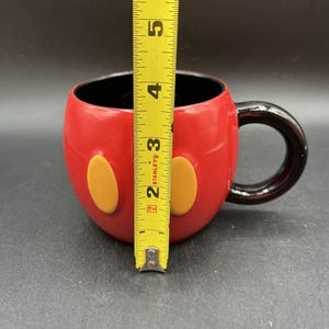 Pu&ograve; includere: Tazza in ceramica rossa con manico e interno neri, con due accenti ovali gialli. La tazza &egrave; alta circa 10 cm, simile a un personaggio dei cartoni animati.