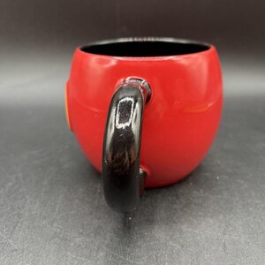 Pu&ograve; includere: Una tazza in ceramica rossa con manico nero. La tazza ha una forma arrotondata e un interno nero. Un piccolo dettaglio giallo &egrave; visibile sul lato della tazza.