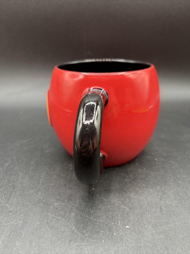 Pu&ograve; includere: Una tazza in ceramica rossa con manico nero. La tazza ha una forma arrotondata e un interno nero. Un piccolo dettaglio giallo &egrave; visibile sul lato della tazza.