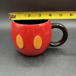Pu&ograve; includere: Tazza in ceramica rossa con interno e manico neri, con due accenti ovali gialli. La tazza &egrave; larga circa 10 cm. Un metro a nastro giallo e nero &egrave; posto sopra la tazza.