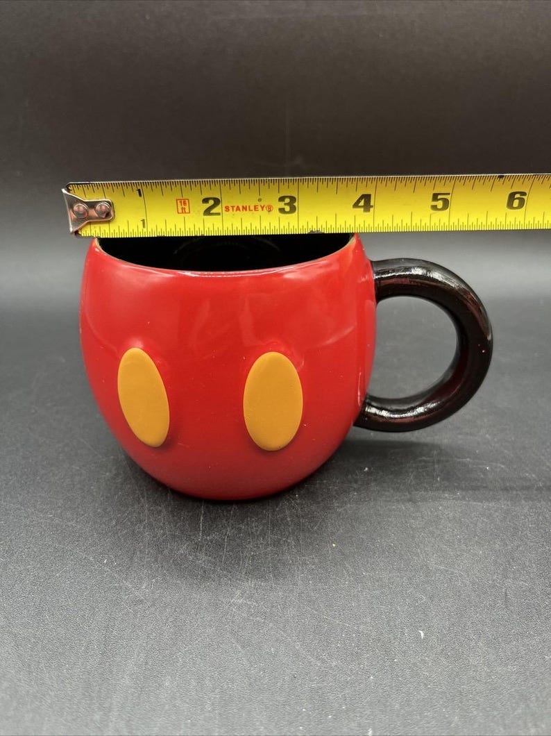 Pu&ograve; includere: Tazza in ceramica rossa con interno e manico neri, con due accenti ovali gialli. La tazza &egrave; larga circa 10 cm. Un metro a nastro giallo e nero &egrave; posto sopra la tazza.