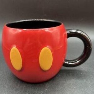 Pu&ograve; includere: Una tazza in ceramica rossa con manico e interno neri. La tazza presenta due forme ovali gialle sul davanti, che ricordano i pantaloncini di Topolino. La tazza &egrave; progettata per assomigliare al volto di Topolino.