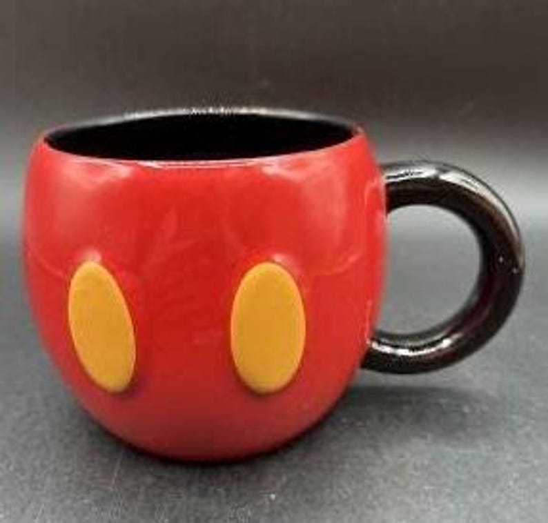 Pu&ograve; includere: Una tazza in ceramica rossa con manico e interno neri. La tazza presenta due forme ovali gialle sul davanti, che ricordano i pantaloncini di Topolino. La tazza &egrave; progettata per assomigliare al volto di Topolino.
